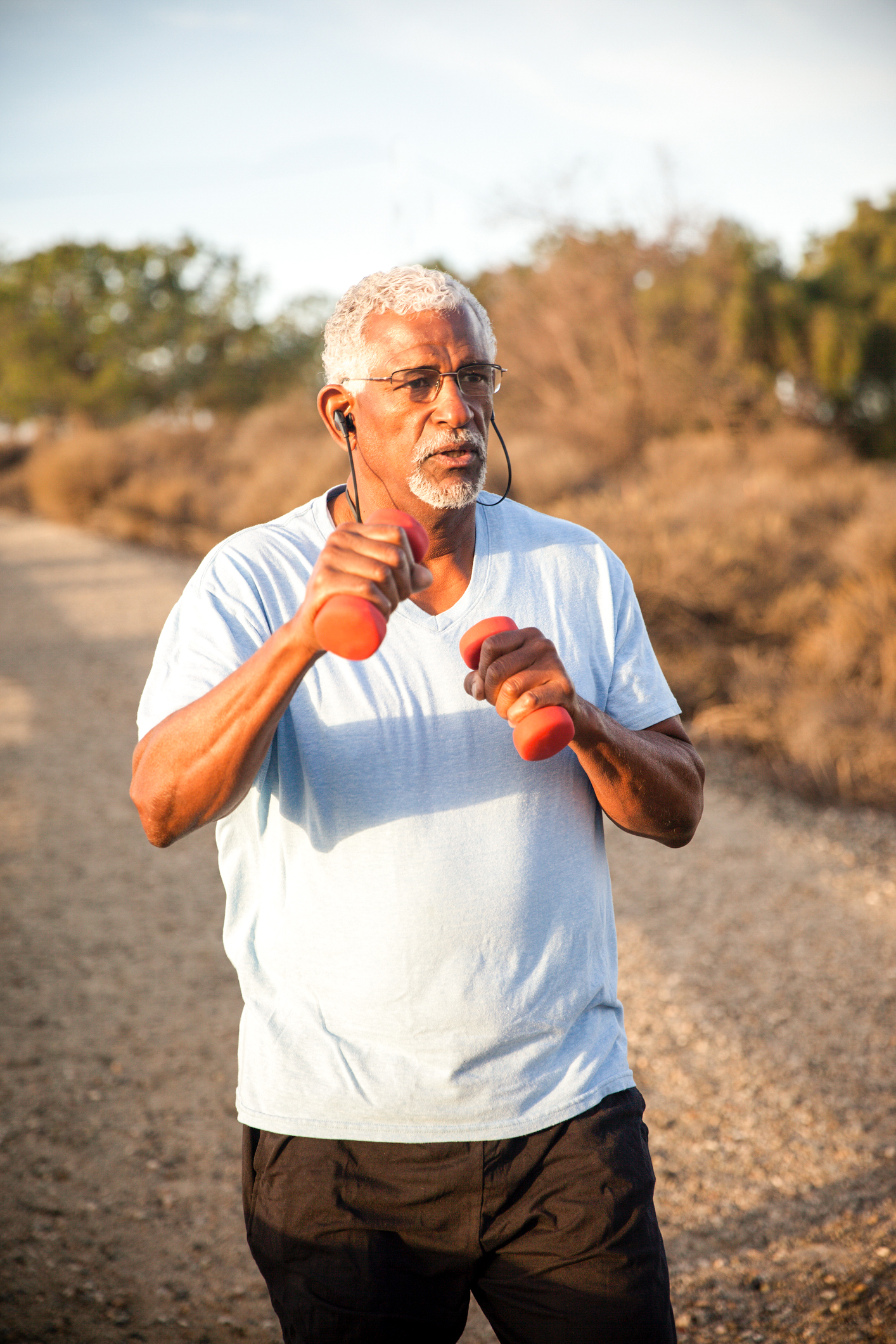 A person with dark skin tone, white hair, and a goatee, wearing a blue shirt, working out outdoors with hand weights.